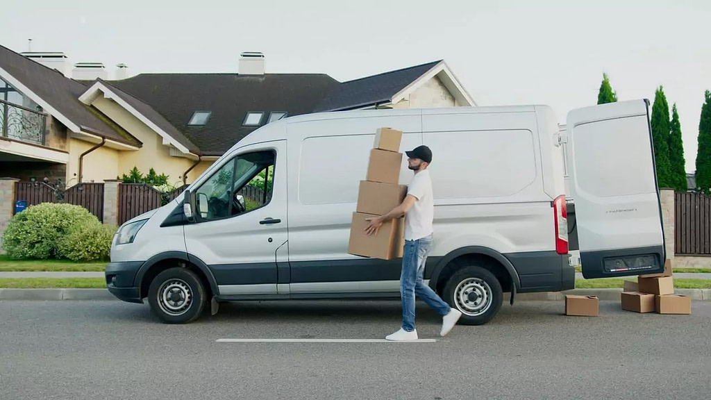 Corriere consegna una pila di pacchi, davanti a un furgone bianco con portellone aperto in un quartiere residenziale